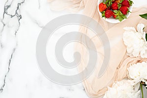 Top view pastel beige scarf, bowl of strawberry and white peony flowers on marble background. Minimal flat lay style composition