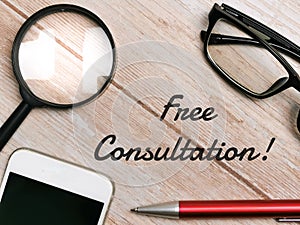 Top view pen,eye glasses,smartphone and magnifying glass with text Free Consultation written on wooden background.