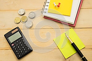 top view. pen putting on yellow post-it note and have black calculator, coin, pin and more post-it note putting on notebook. wood