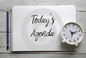 Top view of pencil,clock and notebook written with Today`s Agenda on white wooden background