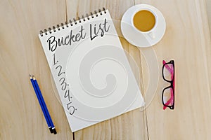 Top view of pencil,glasses,a cup of coffee and notebook written with Bucket List on wooden background.