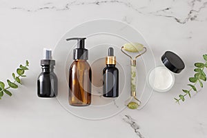 Top view photo of amber cosmetic bottles, cream jar, dropper bottle, face massage roller and eucalyptus leaves