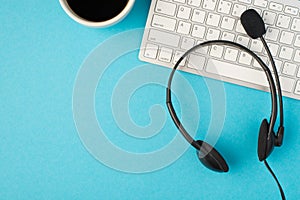 Top view photo of black headphones with microphone on white keyboard and cup of coffee on isolated light blue background with