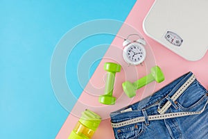 Top view photo of blue jeans with tape measure,  dumbbells, scales and alarm clock on pink and blue background