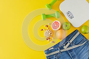 Top view photo of blue jeans with tape measure,  dumbbells, scales and fruit on yellow background
