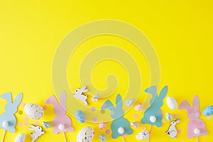Top view photo of color easter eggs, cute rabbits, confetti and paper bunny toppers on yellow background