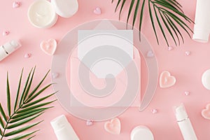 Top view photo of cosmetic bottles, green tropical leaves, heart shaped candles on pastel pink background