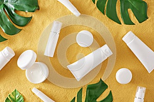 Top view photo of cosmetic bottles without label, cream jars and green tropical leaves on sandy background
