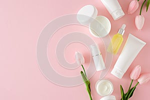 Top view photo of cosmetic tubes without label, cream jars, serum bottles and spring flowers on pastel pink background