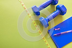 Top view photo of dumbbells, measuring tape and notepad on a green background with blank space