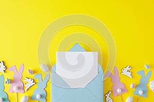 Top view photo of envelope with card, cute rabbits, multicolored eggs, paper bunny toppers on yellow background