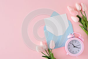 Top view photo of envelope with letter, spring flowers and alarm clock on pastel pink background