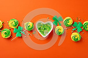 Top view photo of heart shaped saucer with sprinkles meringue lollipops and shamrocks on isolated vibrant orange background