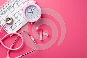 Top view photo of pink ribbon symbol of breast cancer awareness alarm clock calendar and stethoscope on isolated pink background