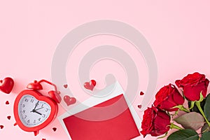 Top view photo of red flowers, heart shaped alarm clock, envelope, hearts on pastel pink background