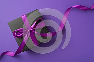 Top view photo of stylish black giftbox with violet satin ribbon bow on isolated violet background with blank space