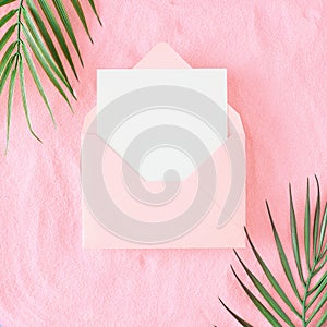 Top view photo of tropical leaves on pink sandy background and open envelope with card