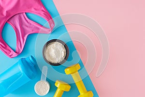 Top view photo of whey protein in jar with shaker, pink sports top and dumbbells on pastel pink and blue background