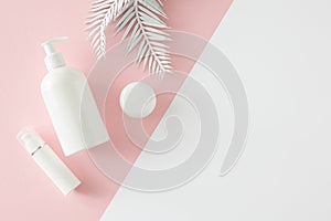 Top view photo of white cosmetic bottle, cream jars, spray bottle and tropical leaves on pastel pink and white background