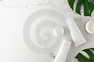 Top view photo of white cosmetic tubes, cream jars and green tropical leaves on marble background