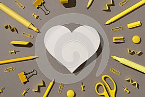 Top view photo of white paper heart surrounded by yellow stationery school supplies felt pens pencils pins binder clips magnets