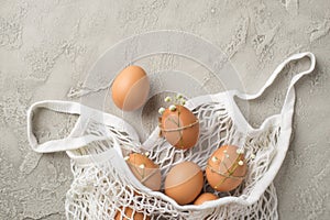 Top view photo of white string bag with brown eggs decorated by gypsophila flowers and twine on isolated grey concrete background