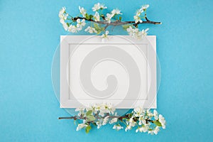 Top view photo of white wooden photo frame and branch with flowers on pastel blue background