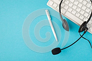 Top view photo of wired black headset pen and white keyboard on isolated pastel blue background with blank space