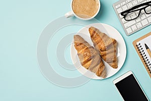 Top view photo of workplace keyboard glasses notebook pen mobile phone cup of frothy coffee and plate with two croissants on