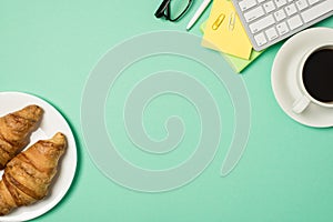 Top view photo of workplace keyboard sticky note paper clips pen glasses croissants on plate and cup of coffee on saucer on