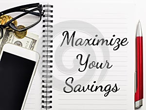 Top view phrase Maximize your savings written on note book with pen,fake money,coins,smartphone and eye glasses.
