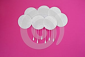 Top view on pink cotton buds with white heads and white round cotton disks laid out in clouds with rain drops on a pink