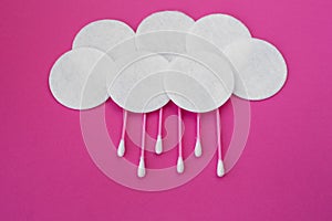 Top view on pink cotton buds with white heads and white round cotton disks laid out in clouds with rain drops on a pink background