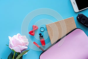 Top view of pink lady bag with diary book, pen, lipstick, bow, ring, car key, smartphone and sweet rose on blue background