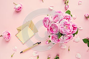 Top view pink tea roses bouquet,blank of greeting card and craft paper envelope, pen on pink background. Postcard mock up. Summer,