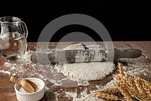 Top view of pizza dough with rolling pin, on wooden table with flour, jug of water, salt and wheat, selective focus, black backgro