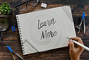 Top view of plant,sunglasses,magnifying glass,stationery and hand holding pen writing Learn More on notebook