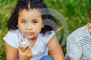 Top view portrait of cute hispanic dark skinned curly sweet girl eating ice cream, sitting on the green grass at park outdoor.