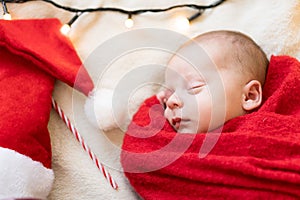 Top View Portrait First Days Of Life Newborn Cute Sleeping Baby In Santa Hat Wrapped In Red Diaper rug At White Garland