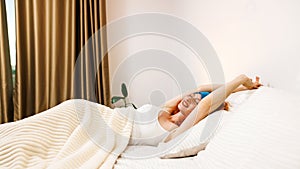 Top view portrait of smiling Caucasian young woman in sleeping mask relaxing on cozy bed at home or hotel, happy