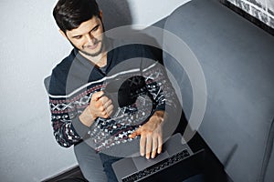 Top view portrait of young confident man with black mug in hand, sitting home on sofa and using laptop.