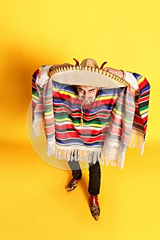 Top view portrait of young man in colorful festive clothes, poncho and sombrero posing against yellow studio background