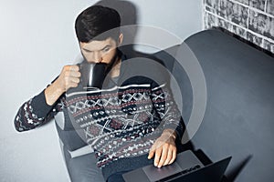 Top view portrait of young man, sitting home on sofa, drinking coffee and looking surprised in laptop.