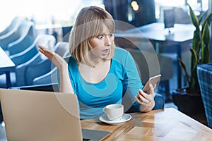 Top view portrait of young shocked blonde woman in blue t-shirt, sitting in cafe and holding her mobile smart phone and looking