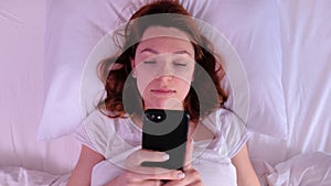 Top view of positive young woman waking up, smiling, stretching in bed and taking her cellphone. Happy girl in white t