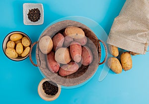top view of potatoes in basket and in bowl with other ones spilling out of sack and black pepper seeds on blue background