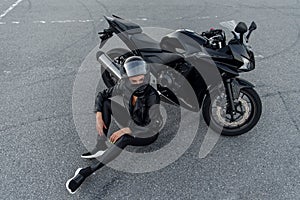 Biker woman in black leather jacket and full face helmet sits near stylish sports motorcycle at urban parking. Traveling