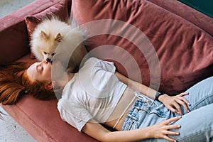 Top view of pretty white small Spitz pet dog lovely licking face of happy young woman lying on comfortable sofa.