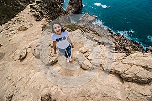 top view of pretty woman in a white t-shirt and blue jeans standing on the rocky edge of the cliff. The waves of the ocean break