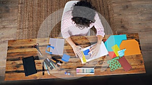 Top view of professional designer working at table making paper collage sticking figures in notebook and cutting shapes